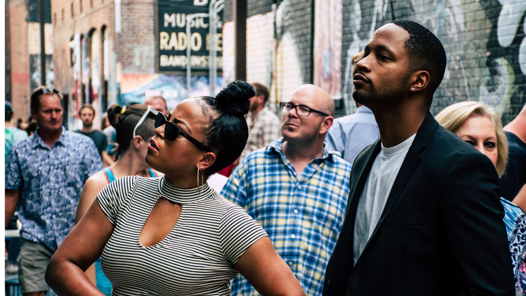 A woman standing looking up on a busy street in a grey shirt and sunglasses and a man standing next to her looking up.