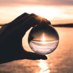 Hand holding a glass see through ball against a sunset over water